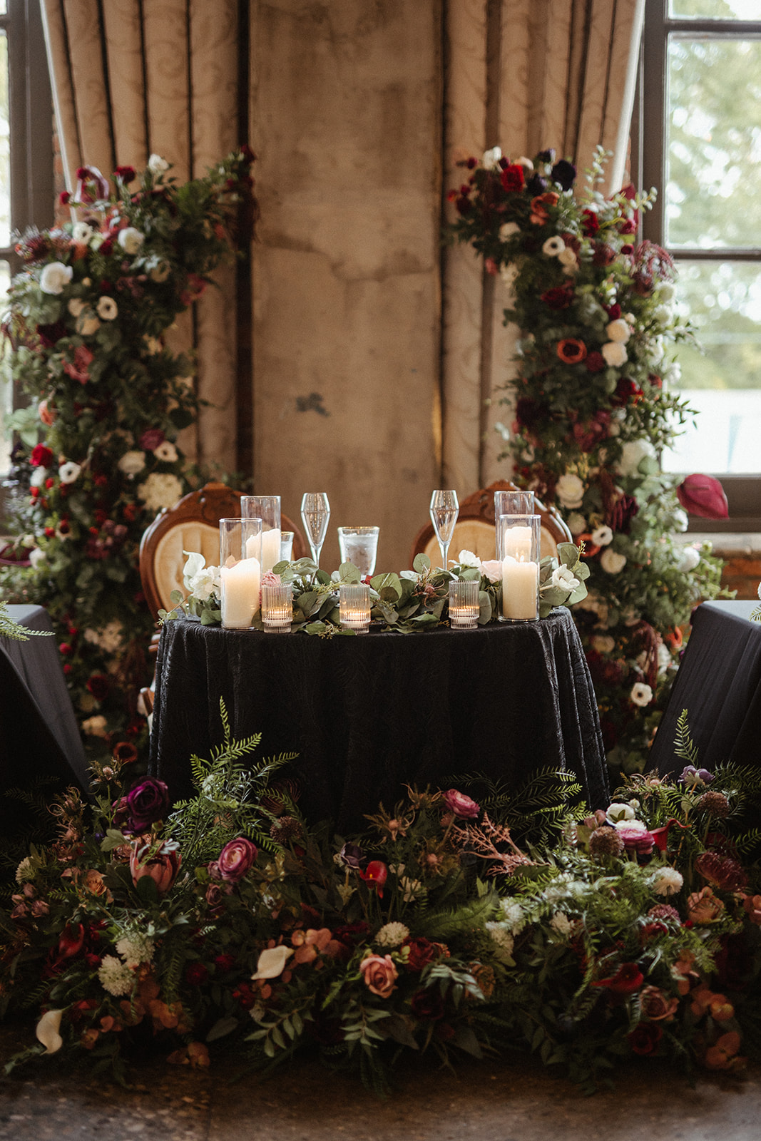 Moody sweetheart table with black velvet linen, pillar candles, eucalyptus garland, and dramatic faux floral columns in burgundy, blush, and white, Steam Plant wedding Dayton Ohio