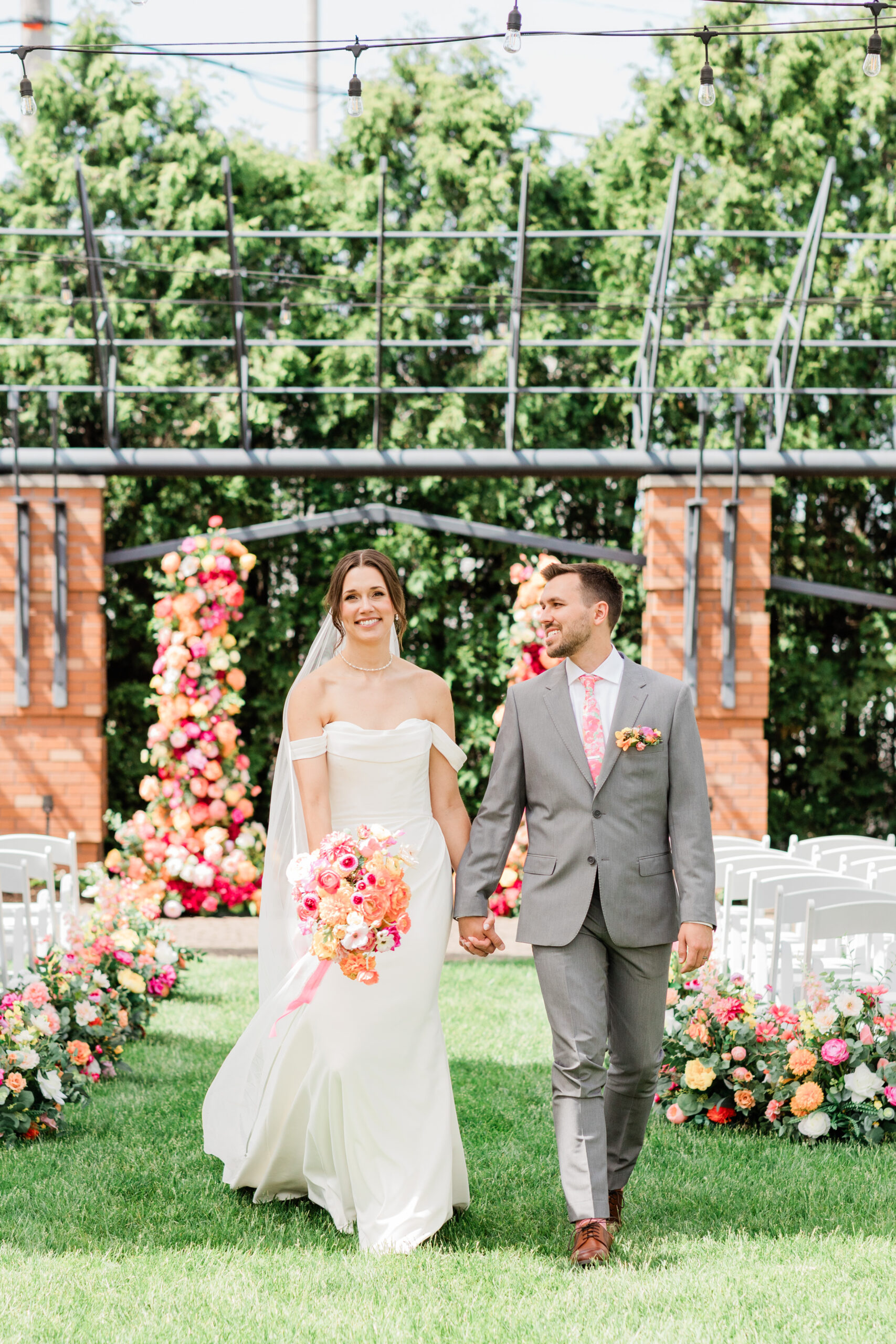 Bride and groom walking down aisle after ceremony with colorful pink and orange faux floral ceremony columns and ground arrangements, outdoor summer wedding