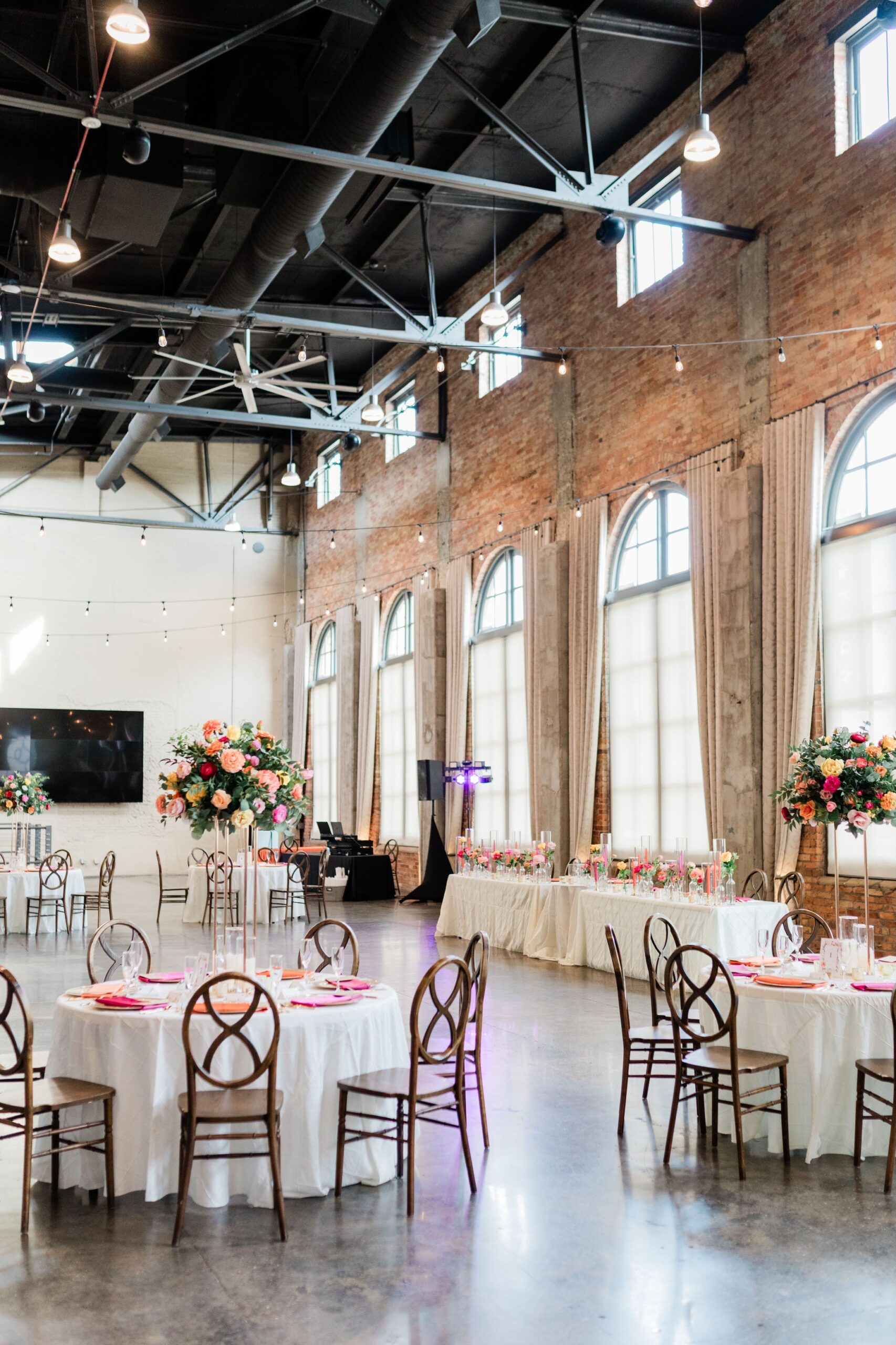 Industrial wedding reception with tall faux floral centerpieces in coral, pink, and orange, exposed brick venue with string lights